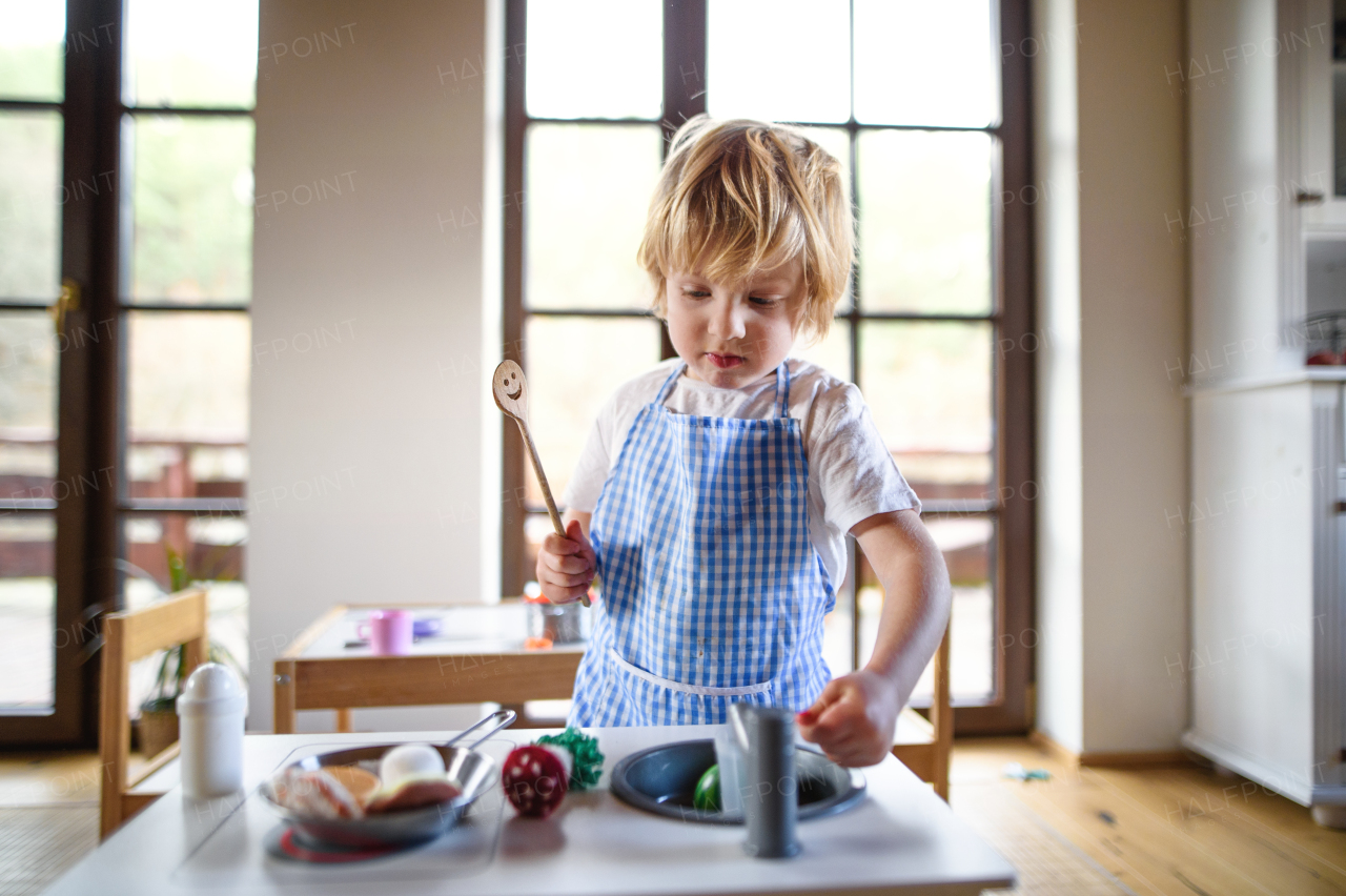 Happy small boy with apron playing indoors with toy kitchen at home.