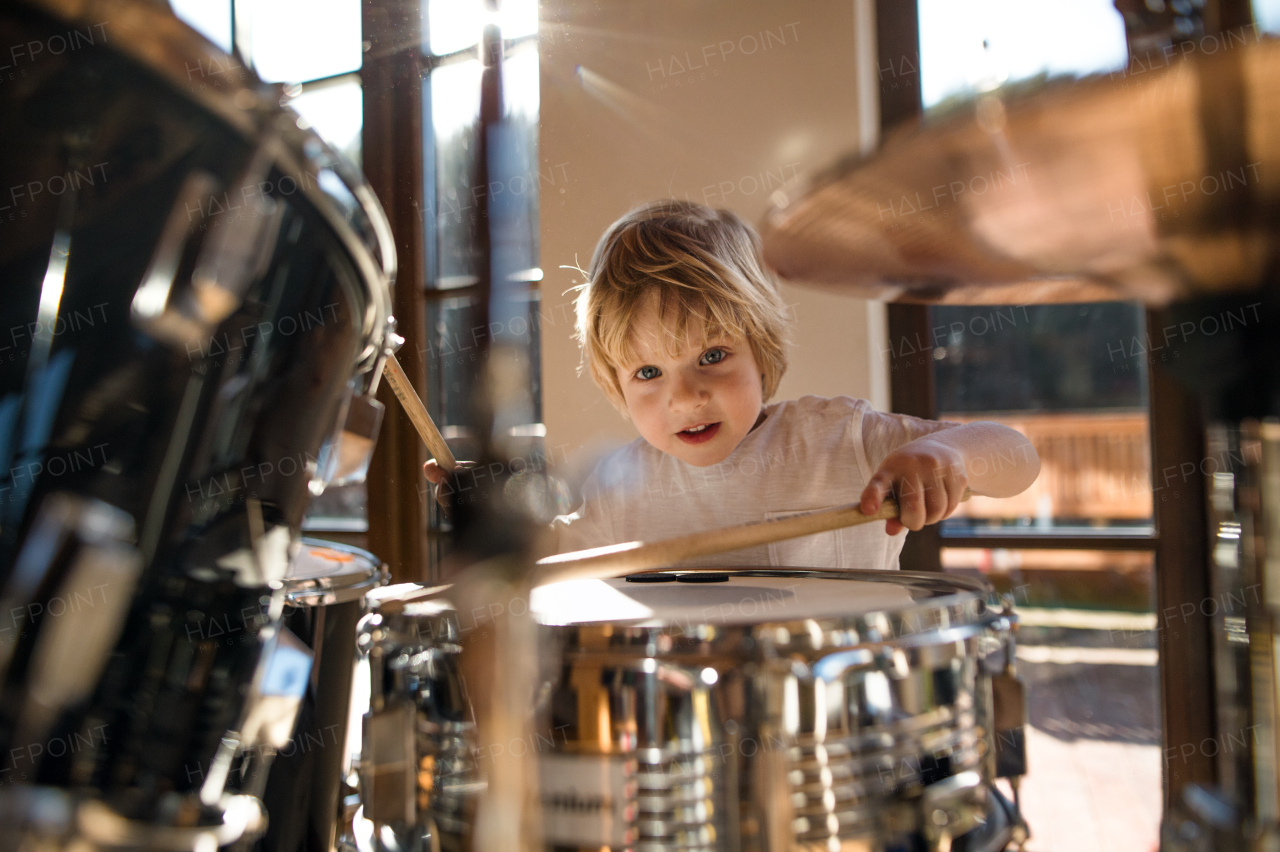 Front view of happy small boy indoors at home, playing drums.