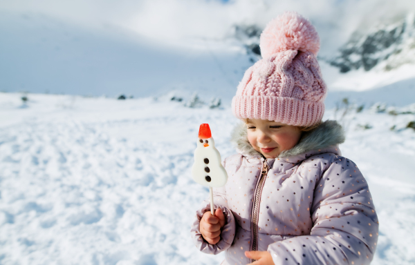 Portrait of cheerful small girl in snow in winter nature, holding snowman lollipop. Copy space.