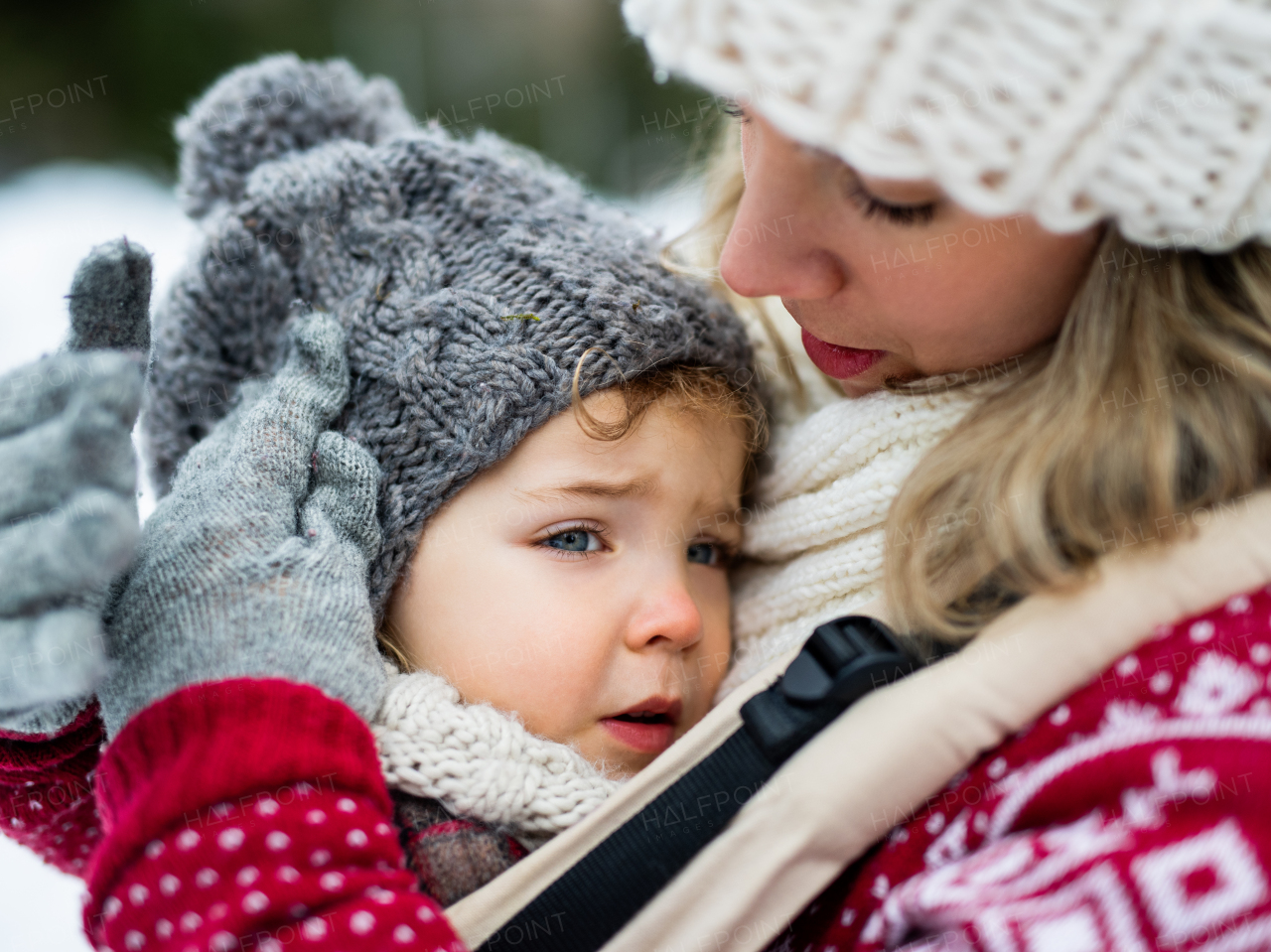 Close-up of upset small daughter in carrier and unrecognizable mother in winter nature.