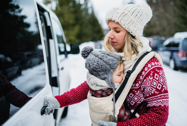 Portrait of mother with sleeping small daughter in carrier standing by car in winter nature.