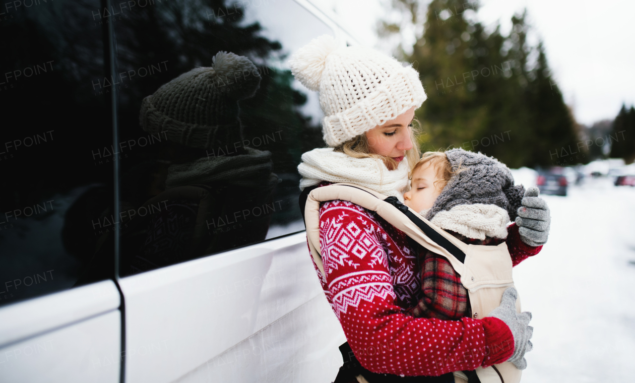 Portrait of mother with sleeping small daughter in carrier standing by car in winter nature.