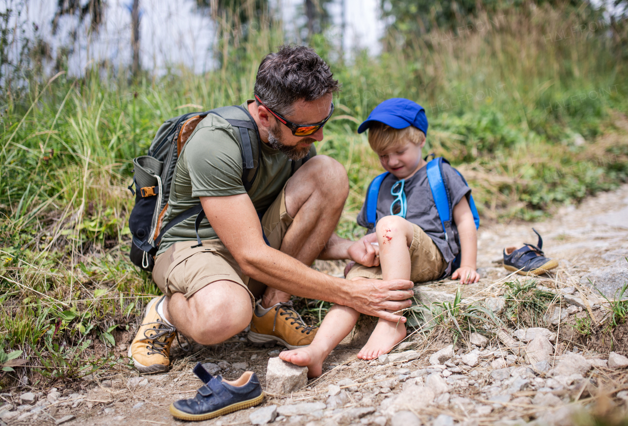 Father with upset small son on trek outdoors in summer nature, falling and scratched knee concept.