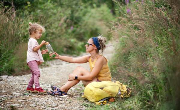 Mother with small toddler daughter hiking outdoors in summer nature, resting and drinking water.