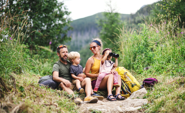 Front view of family with small children hiking outdoors in summer nature, sitting and resting.