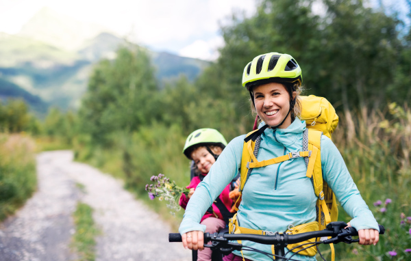 Portrait of mother with small daughter cycling outdoors in summer nature, looking at camera.