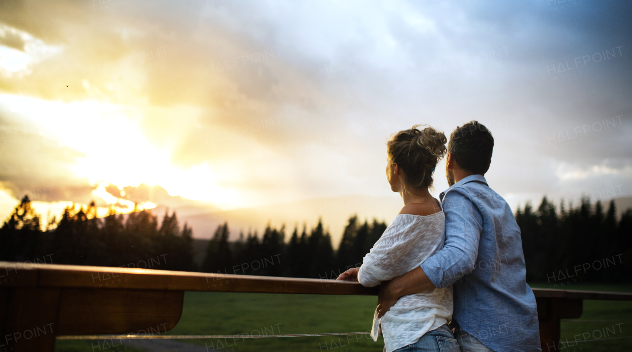 A rear view of couple standing on patio of wooden cabin at sunset, holiday in nature concept.