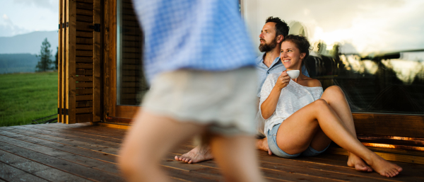 Happy family with small daughter sitting on patio of wooden cabin, holiday in nature concept.