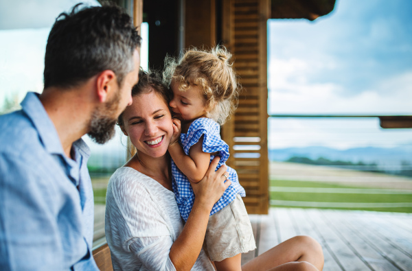 Happy family with small daughter sitting on patio of wooden cabin, holiday in nature concept.