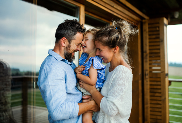 Happy family with small daughter standing on patio of wooden cabin, holiday in nature concept.