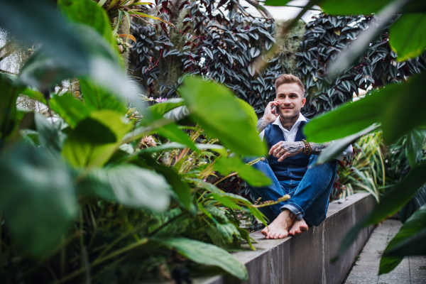 Young man with smartphone sitting in botanical garden, making phone call. Green bussiness concept.