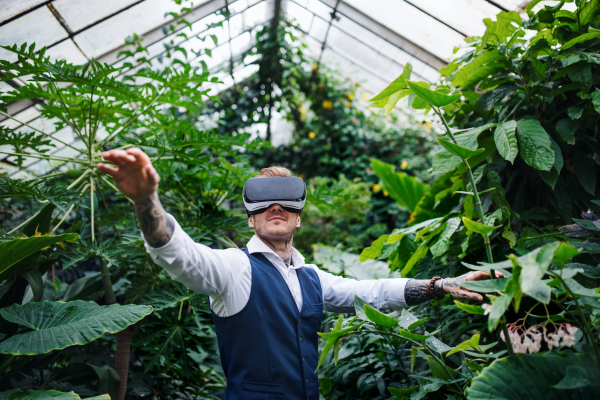 A front view of young man standing in botanical garden, using VR glasses.