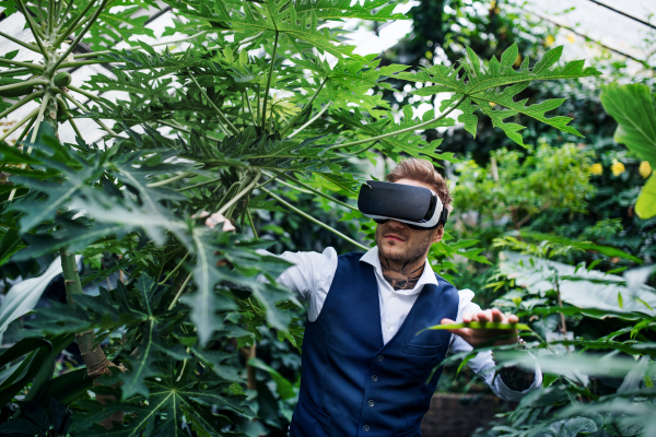 A front view of young man standing in botanical garden, using VR glasses.