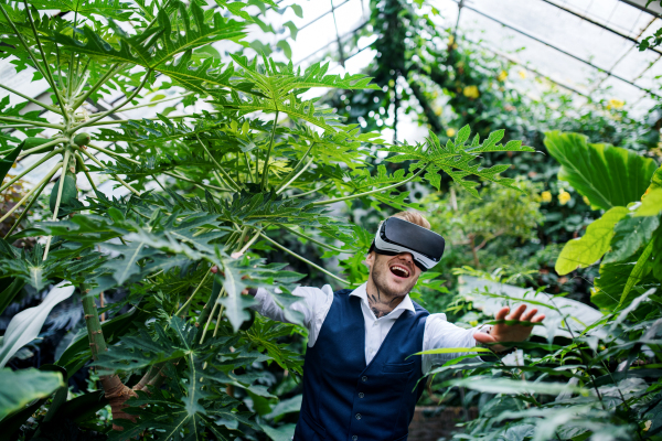 A front view of young man standing in botanical garden, using VR glasses.