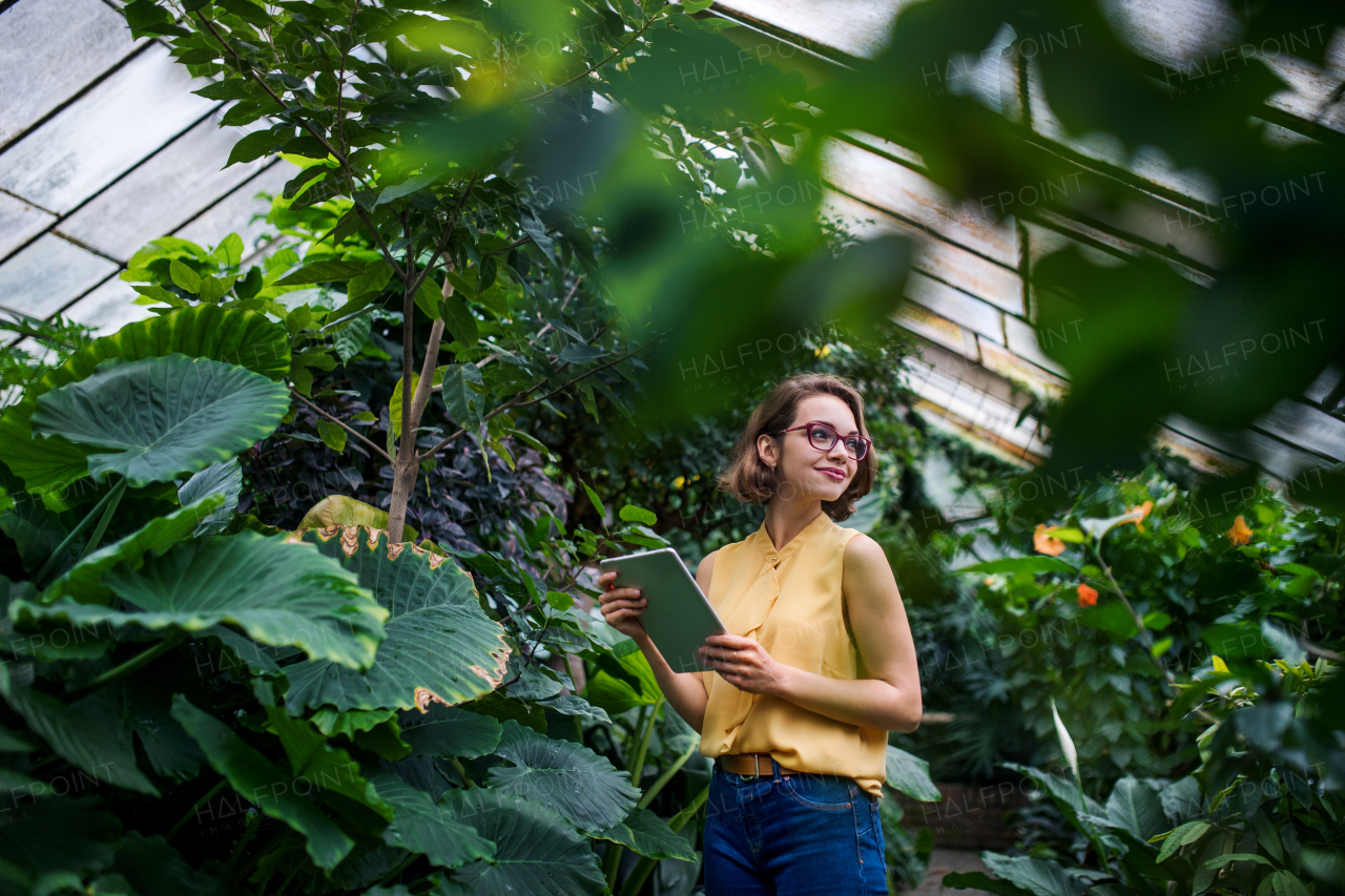 Young woman with tablet standing in greenhouse in botanical garden. Copy space.