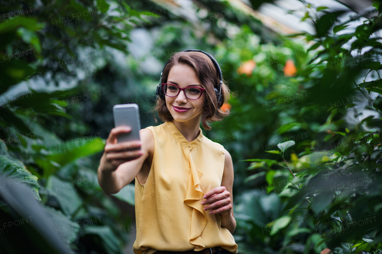 Front view of young woman with smartphone standing in botanical garden, taking selfie.