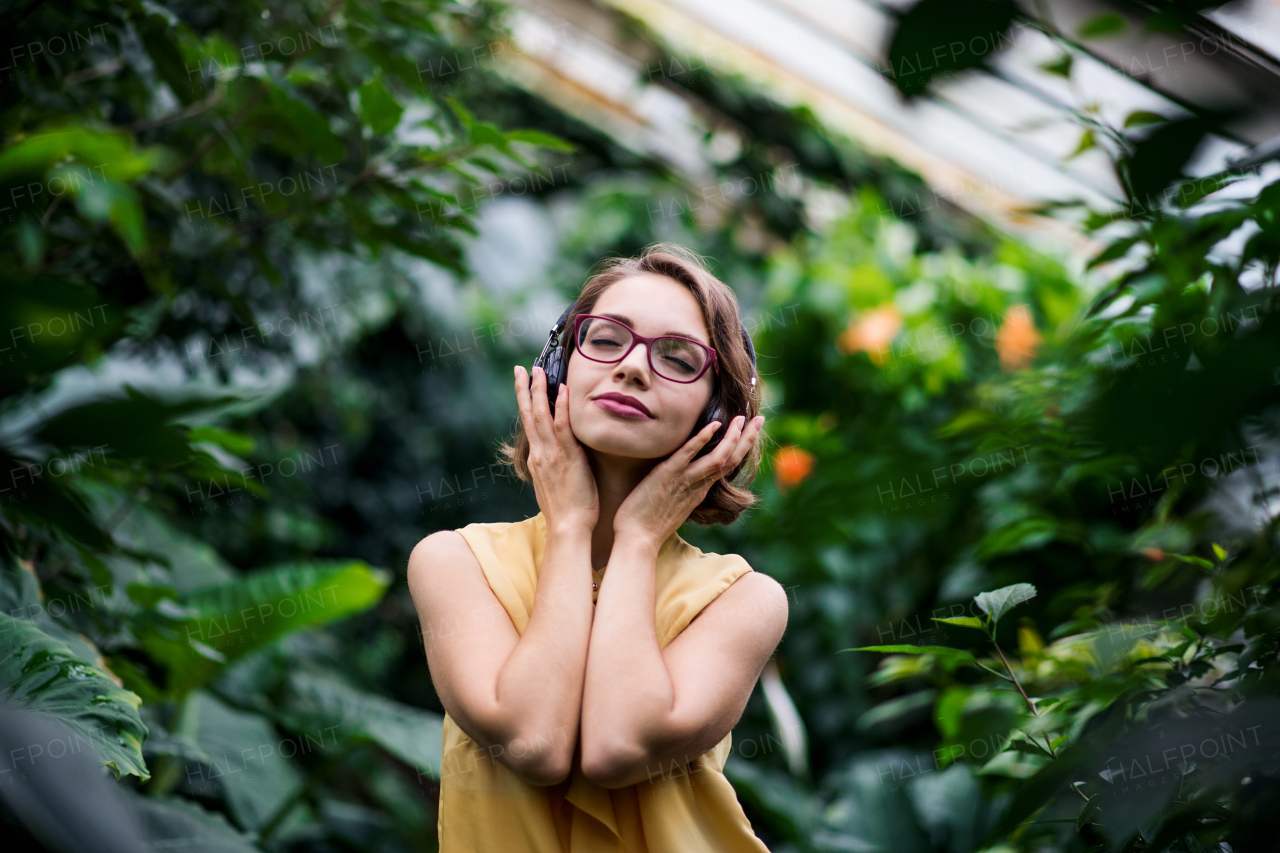 A young woman with headphones standing in greenhouse in botanical garden.