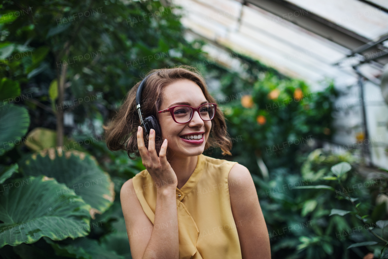 A young woman with headphones standing in greenhouse in botanical garden.