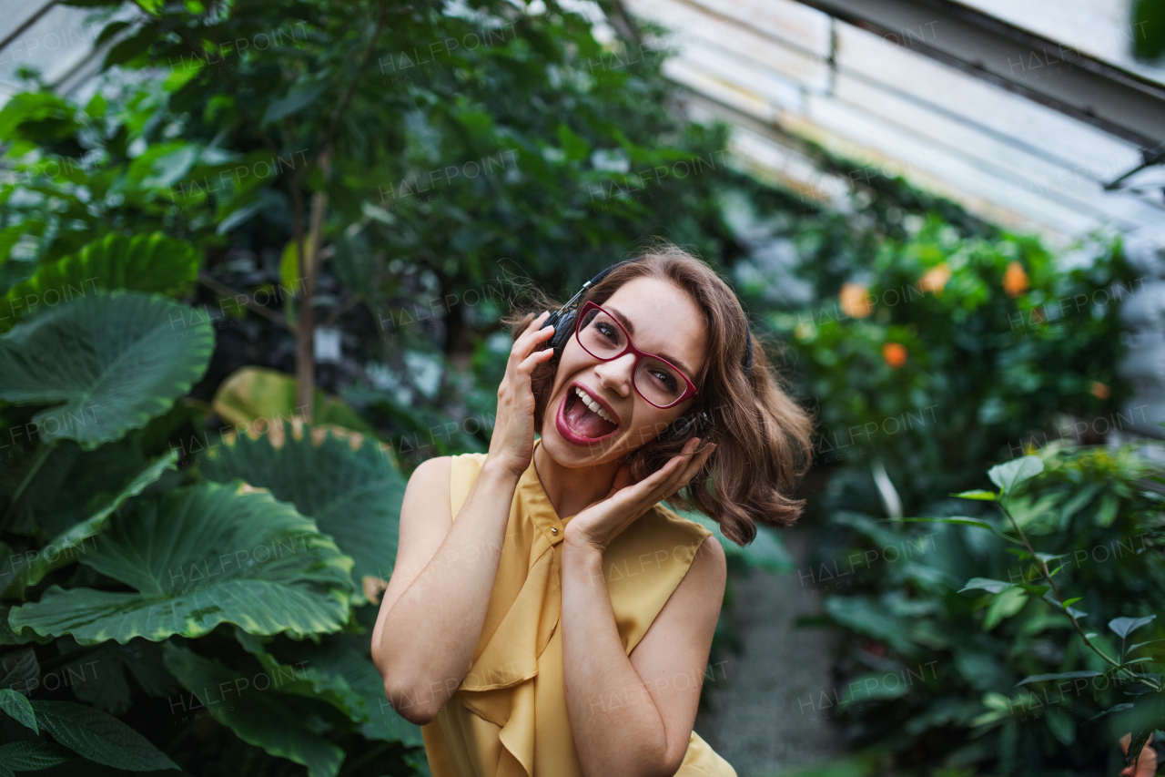 Cheerful young woman with headphones standing in botanical garden, listening to music.