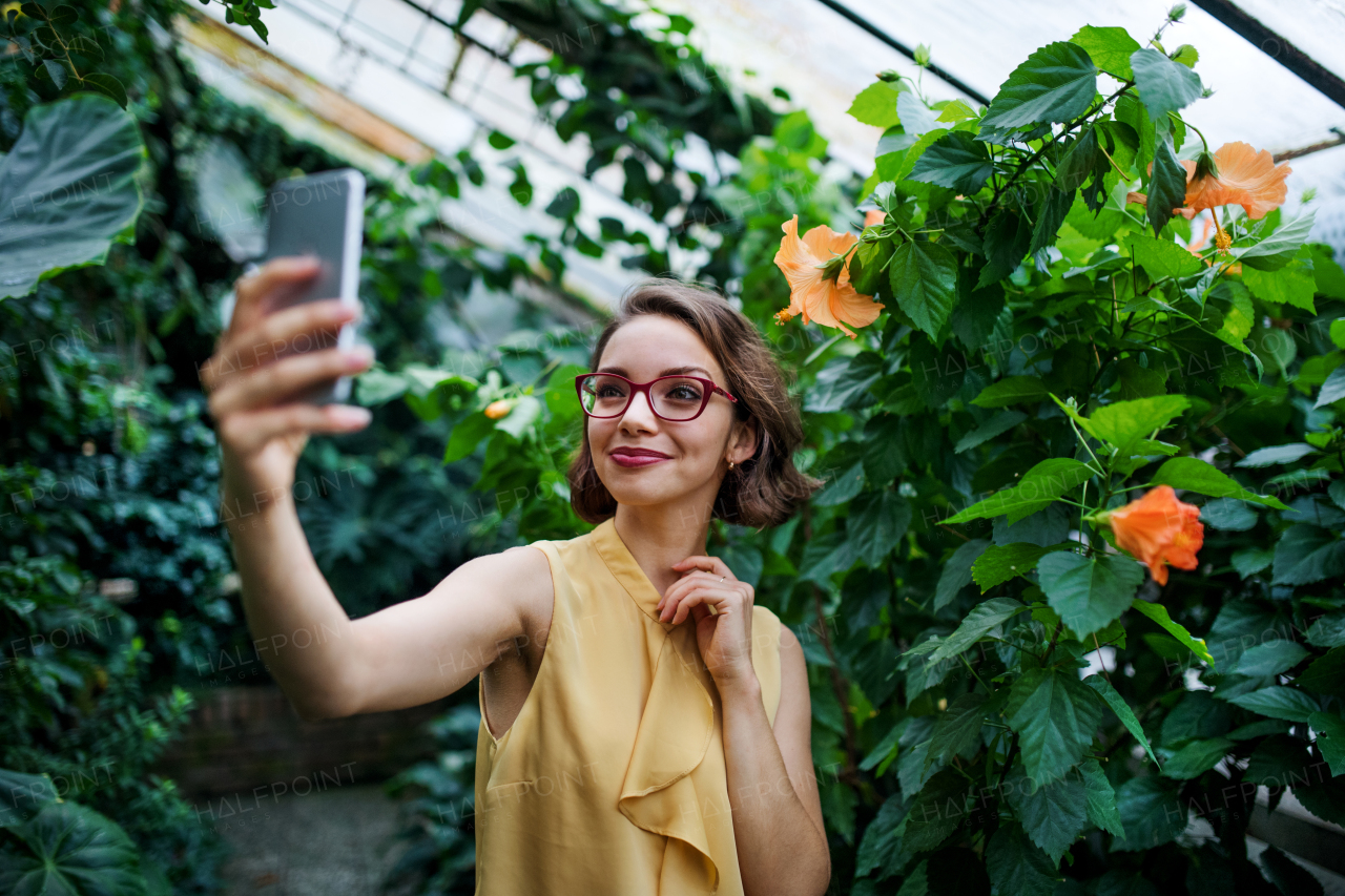 Front view of young woman with smartphone standing in botanical garden, taking selfie.