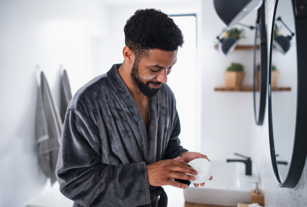 Happy young man applying cream on face ndoors at home, morning or evening routine concept.