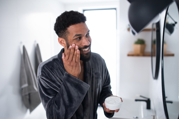 Happy young man applying cream on face ndoors at home, morning or evening routine concept.