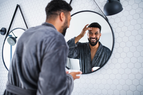 Happy young man applying cream on face ndoors at home, morning or evening routine concept.