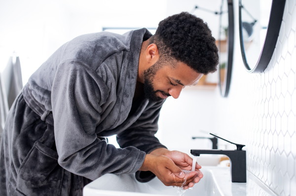 Happy young man washing face ndoors at home, morning or evening routine concept.