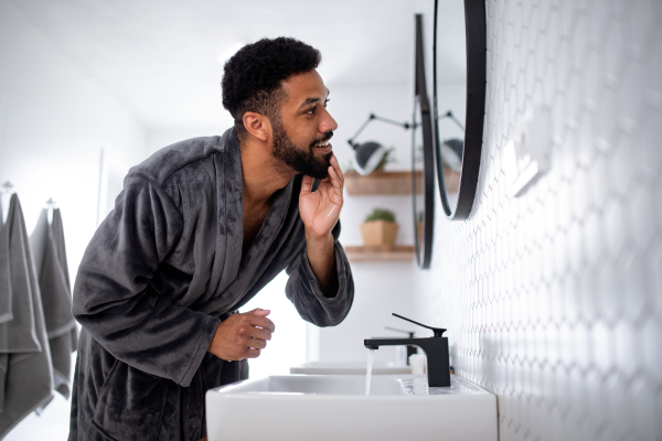 Happy young man washing face ndoors at home, morning or evening routine concept.