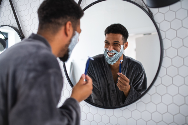 A young man using shaving beard indoors at home, morning or evening routine concept.