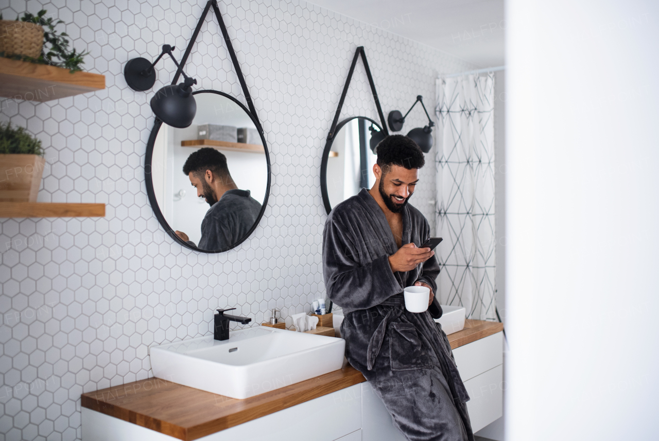 A young man with coffee and bathrobe indoors in bathroom at home, using smartphone.