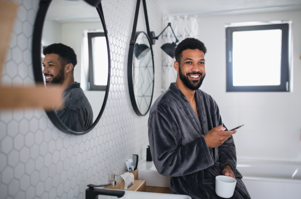 A young man with coffee and bathrobe indoors in bathroom at home, morning routine concept.