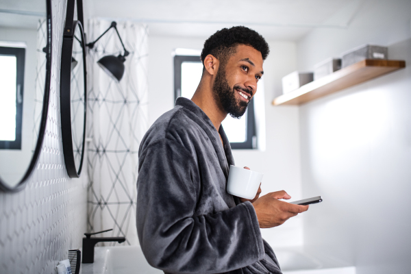 A young man with coffee and bathrobe indoors in bathroom at home, morning routine concept.