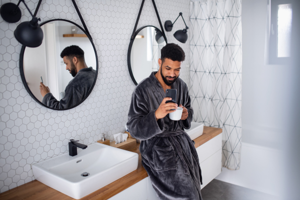 Happy young man with coffee and bathrobe indoors in bathroom at home, taking selfie.