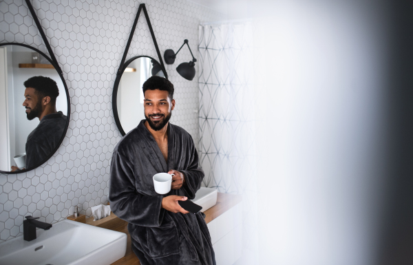 A young man with coffee and bathrobe indoors in bathroom at home, using smartphone.