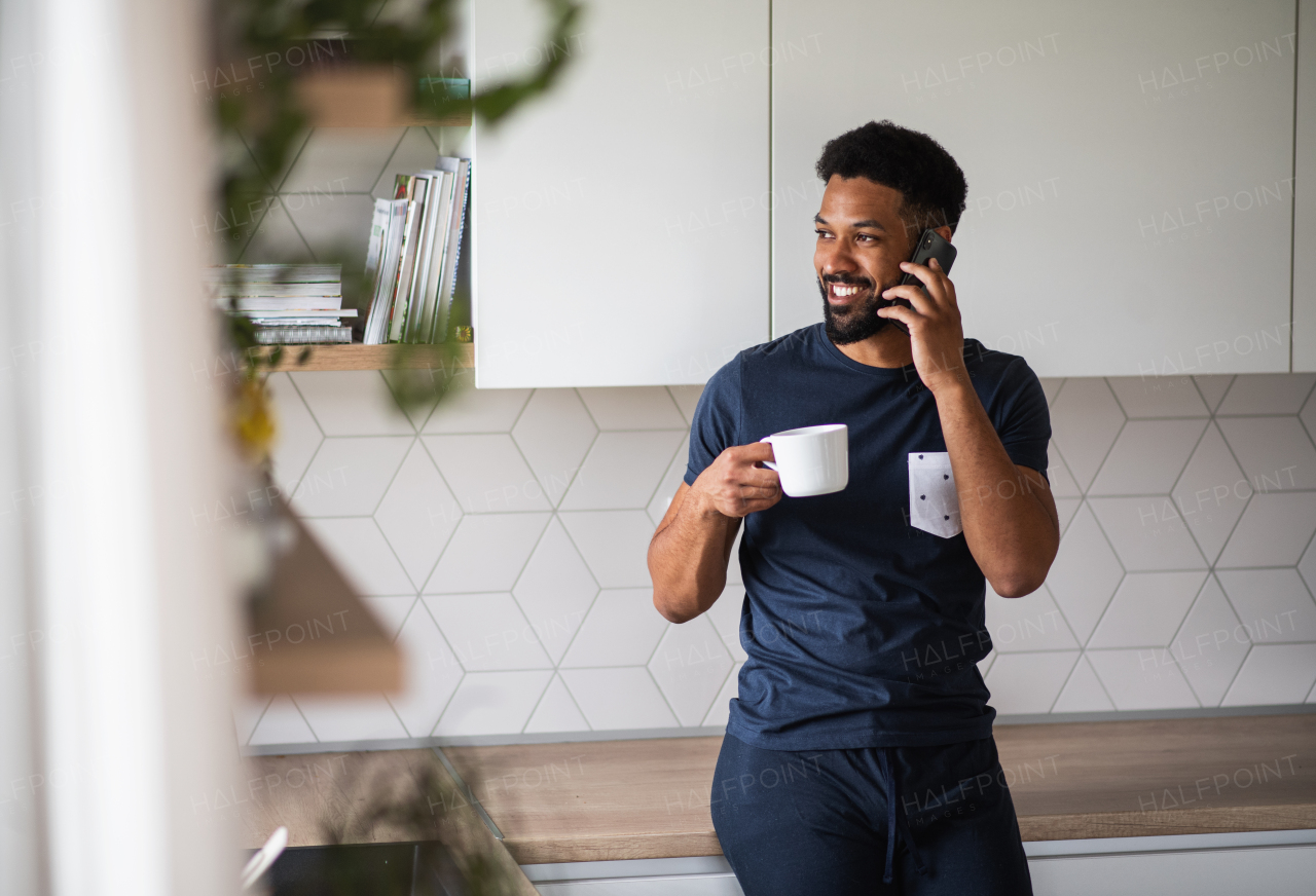 Portrait of young man with cup of coffee standing indoors at home, using smartphone.