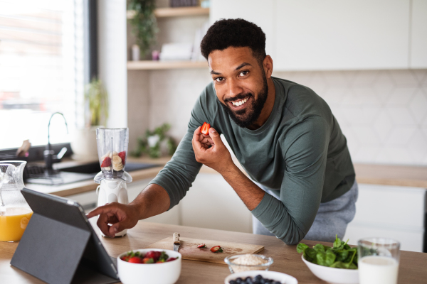 Portrait of young man with tablet preparing healthy breakfast indoors at home, home office concept.