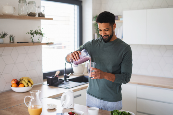 A young man preparing healthy breakfast indoors at home, pouring milk shake into glass.