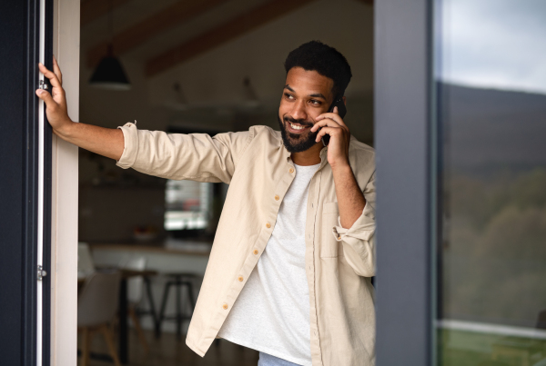 A young man student with smartphone outdoors by patio door at home, making a phone call.