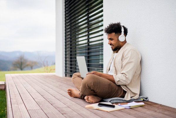 A young man student with laptop and headphones sitting on patio outdoors at home, studying.