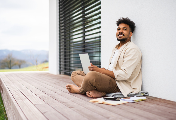 A young man student with laptop sitting on patio outdoors at home, studying.