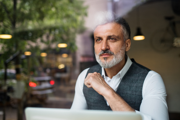 Serious mature man sitting at the table in a cafe, using laptop. Shot through glass.