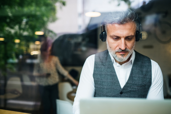 Mature man with headphones at the table in a cafe, using laptop. Shot through glass.