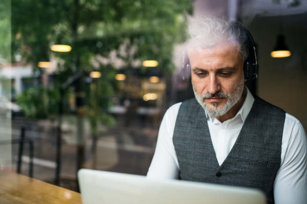 Mature man with headphones at the table in a cafe, using laptop. Shot through glass.