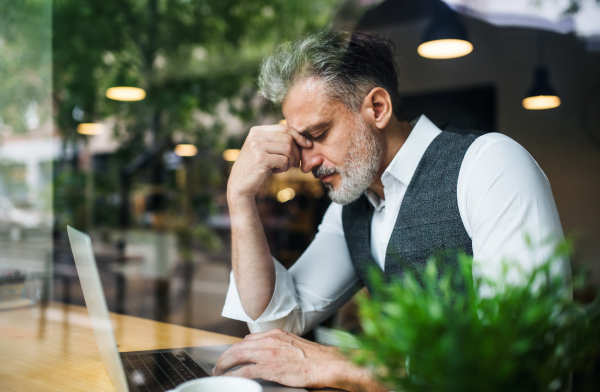 Sad and frustrated mature man sitting at the table in a cafe, using laptop. Shot through glass.