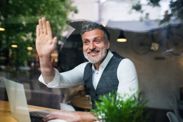 Mature man with laptop at the table in a cafe, greeting somebody. Shot through glass.
