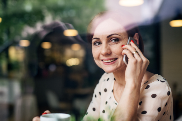 A portrait of woman with coffee and smartphone sitting at the table in a cafe, making phone call. Shot through glass.