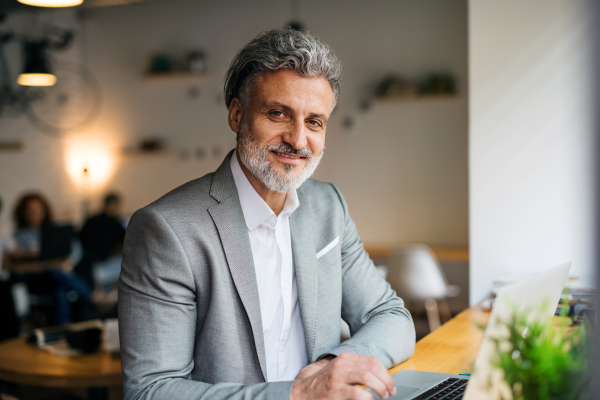 A mature man with laptop at the table in a cafe, looking at camera.
