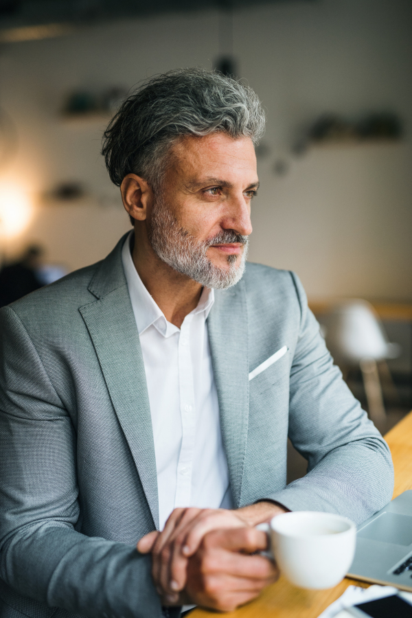 Mature man with coffee and laptop at the table in a cafe, looking out of window.