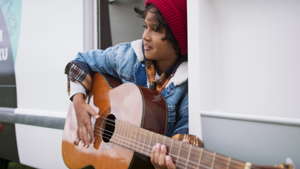 Young multiracial boy in warm clothes playing at guitair during camping at autumn day.